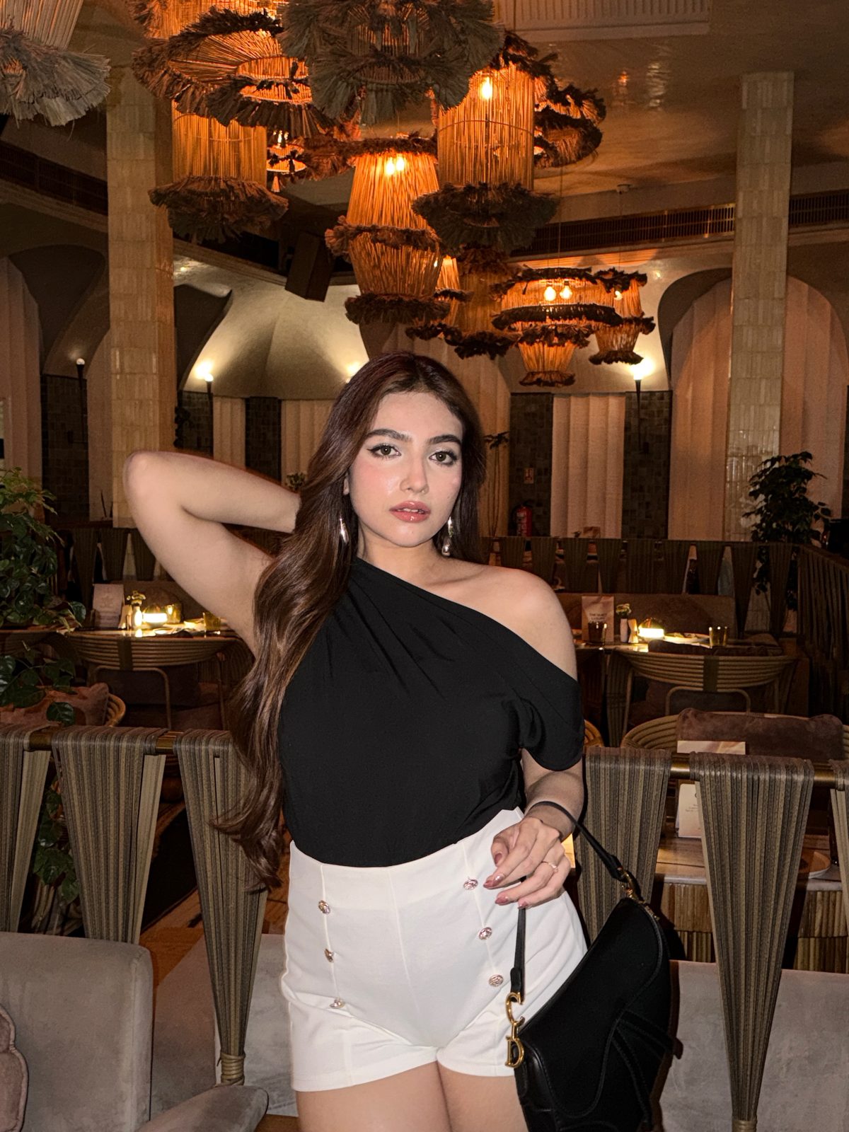 Shrestha Dutta in a black one-shoulder top in a warm, softly-lit restaurant, meeting the camera directly